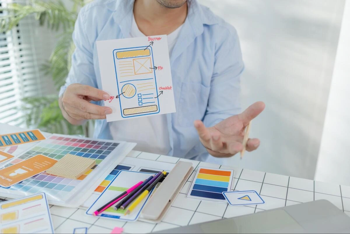 Website Wireframe and SEO Content Planning Web designer holding a hand-drawn website wireframe sketch with color palettes and planning materials on desk