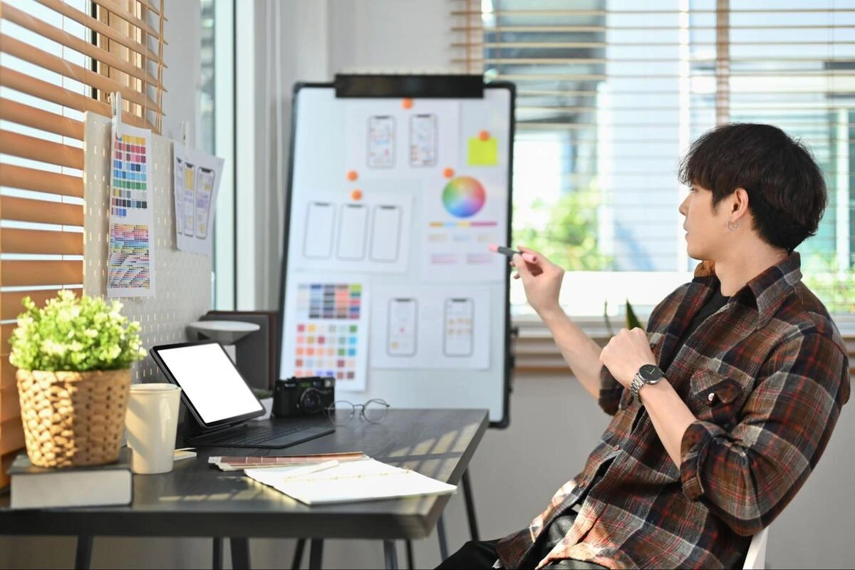 A designer/developer sitting at a desk with a laptop and a drawing table A professional web designer reviewing mobile UI and UX wireframes on a whiteboard.