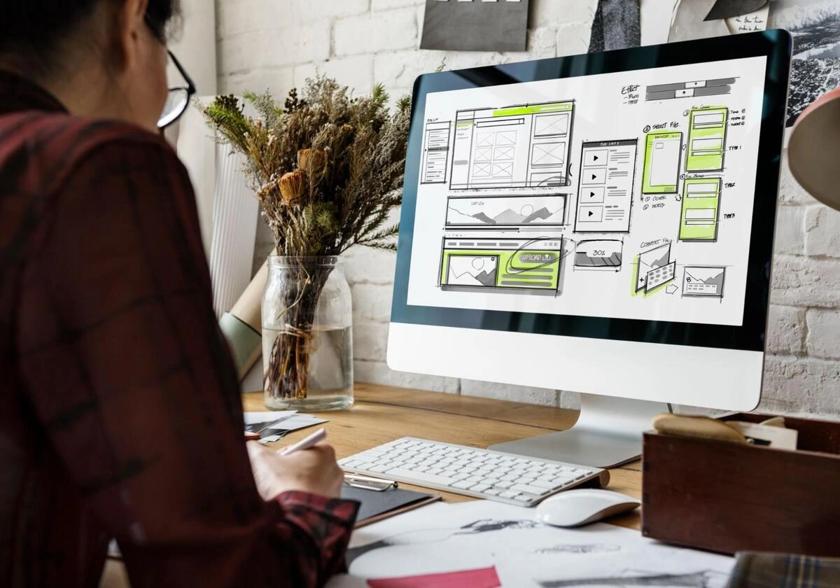 Designer at a desk sketching while a monitor displays website wireframe layouts